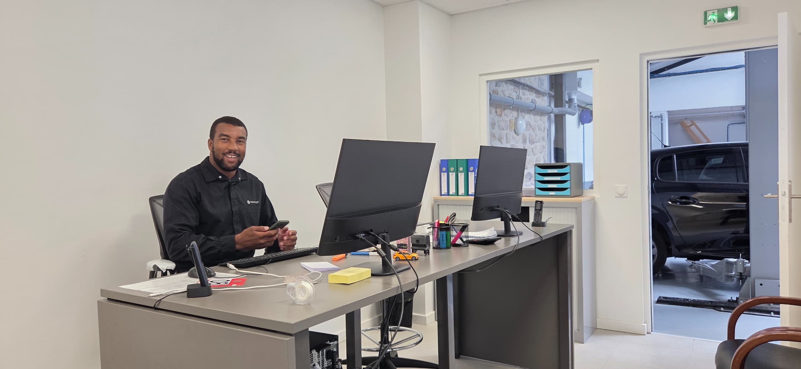 Conseiller souriant au bureau d'accueil du garage à Boulogne-Billancourt, l'atelier et un véhicule sur le pont visibles par la porte